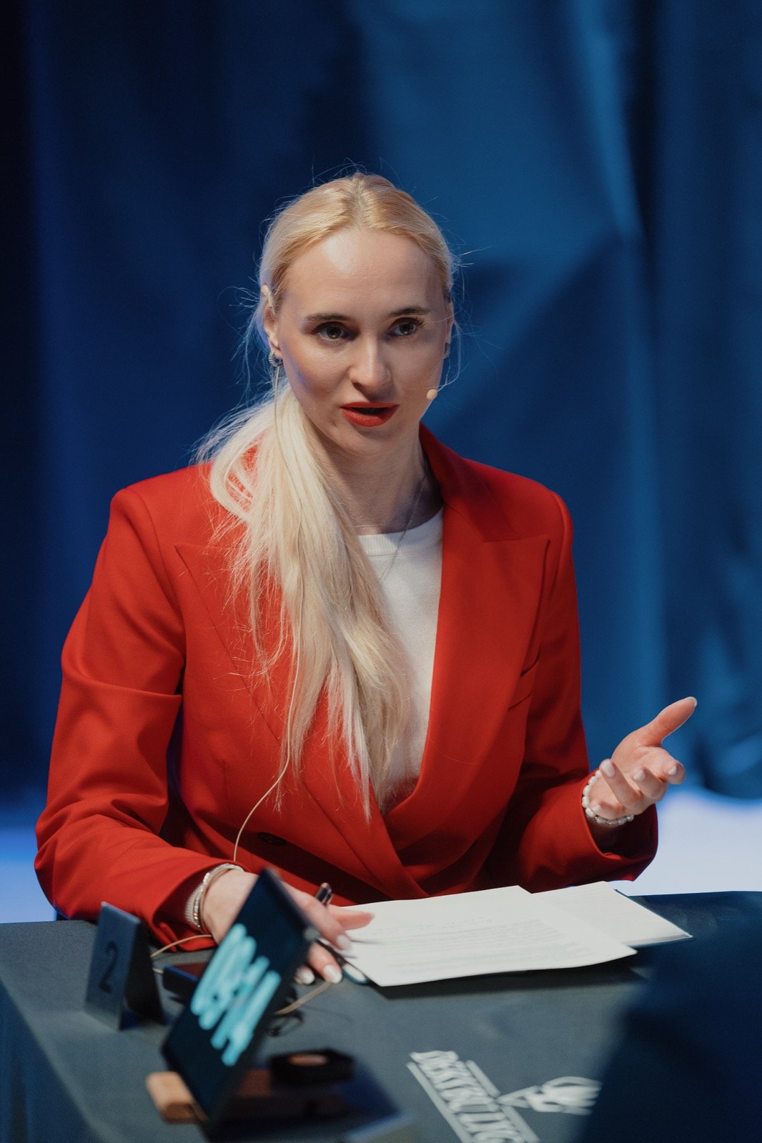 Derybininkė raudonu švarku kalba prie stalo su numeriu 2